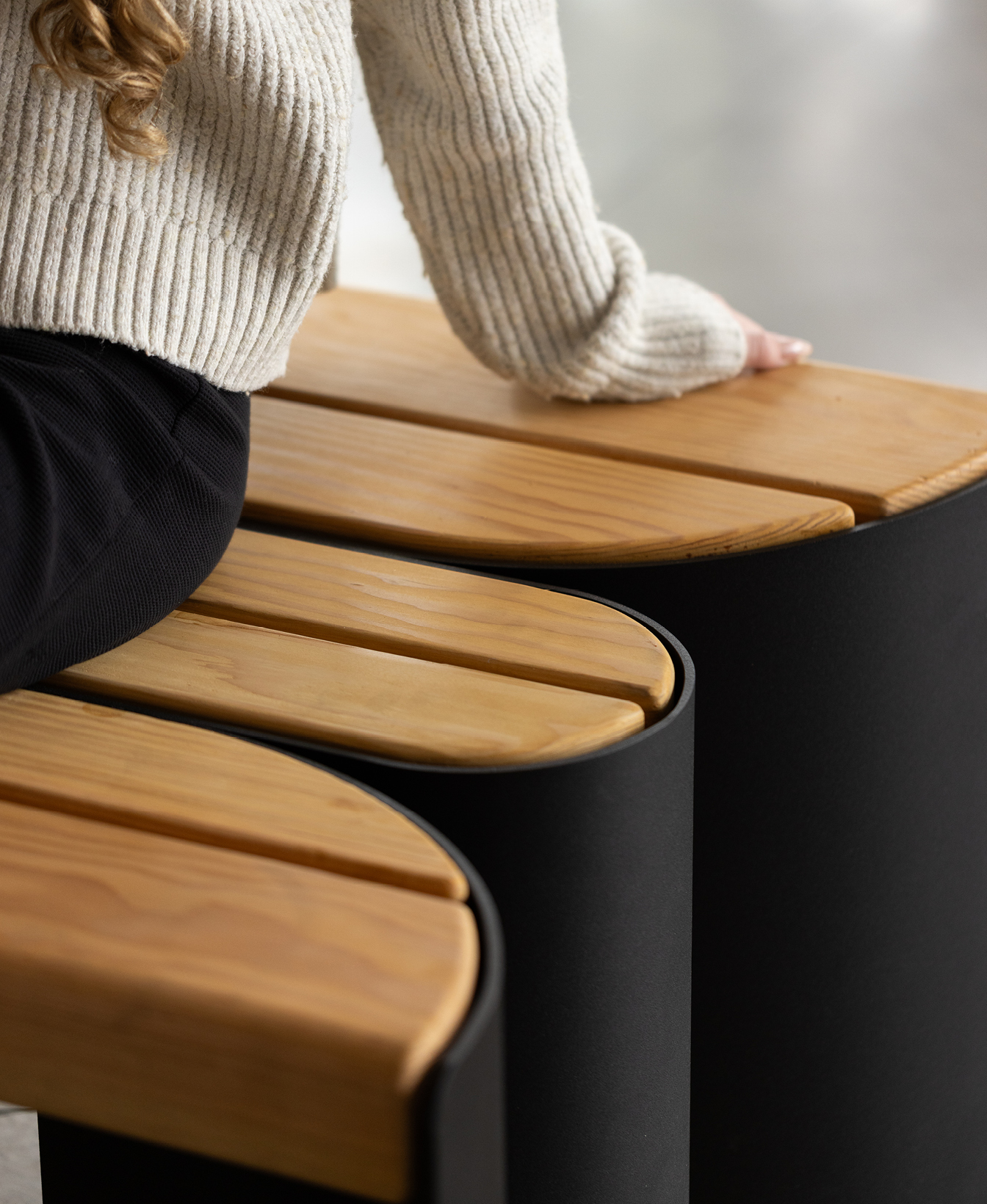 Une personne vêtue d'un pull crème et d'un pantalon sombre est assise sur un banc moderne en bois courbé avec des supports noirs, à l'intérieur.