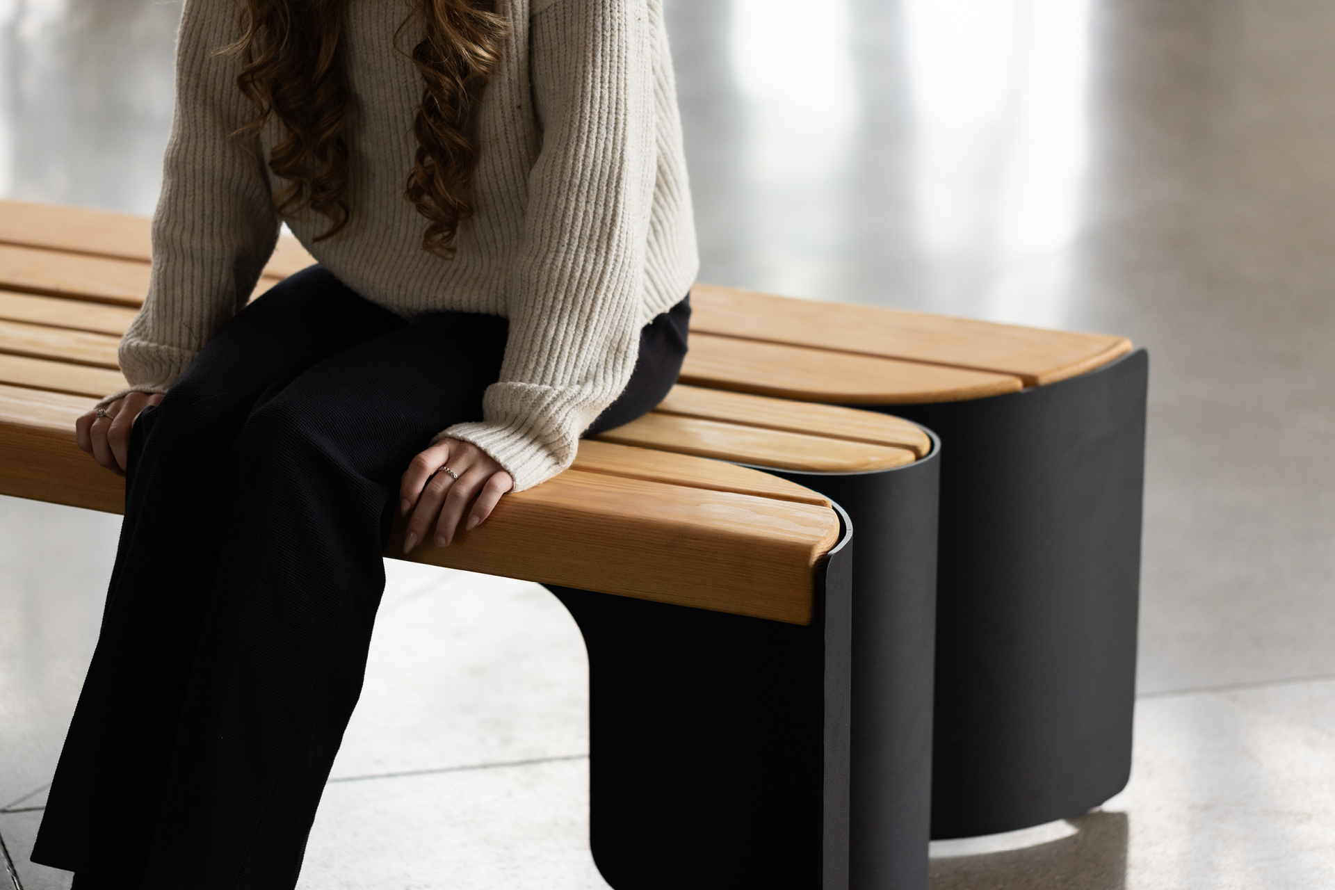 A person with long hair wearing a cream sweater and black pants sits on a modern Maglin wooden bench in a bright indoor space, showcasing stylish site furniture.