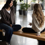 Two women sit on a Maglin wooden bench indoors, facing each other and talking, with plants and large windows in the background, showcasing stylish site furniture.