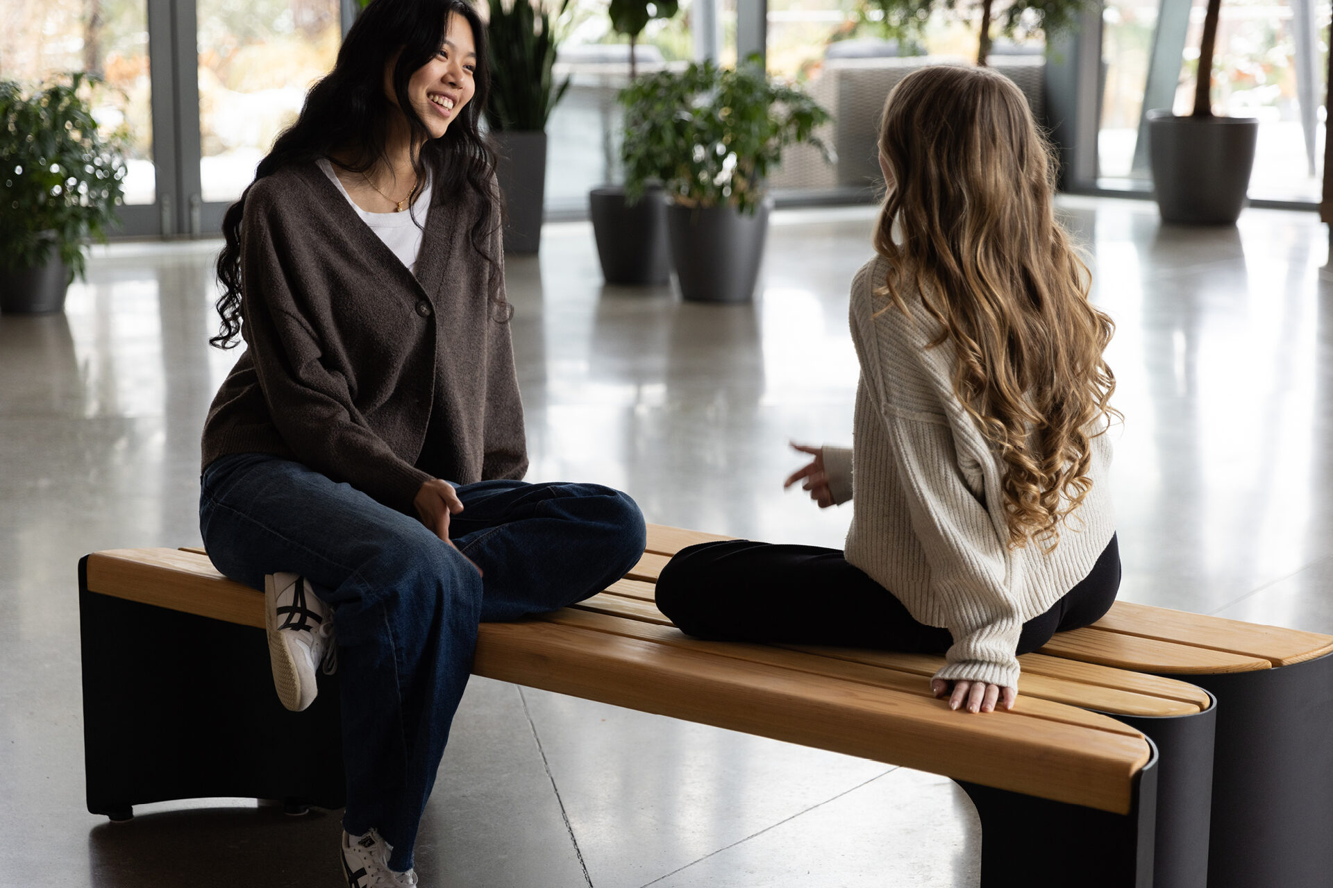 Two women sit on a Maglin wooden bench indoors, facing each other and talking, with plants and large windows in the background, showcasing stylish site furniture.