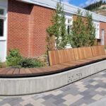 A long wooden Ogden bench with a backrest from the Ogden Collection is built into a curved concrete base in front of a red brick building with windows, small trees, and a patterned walkway.