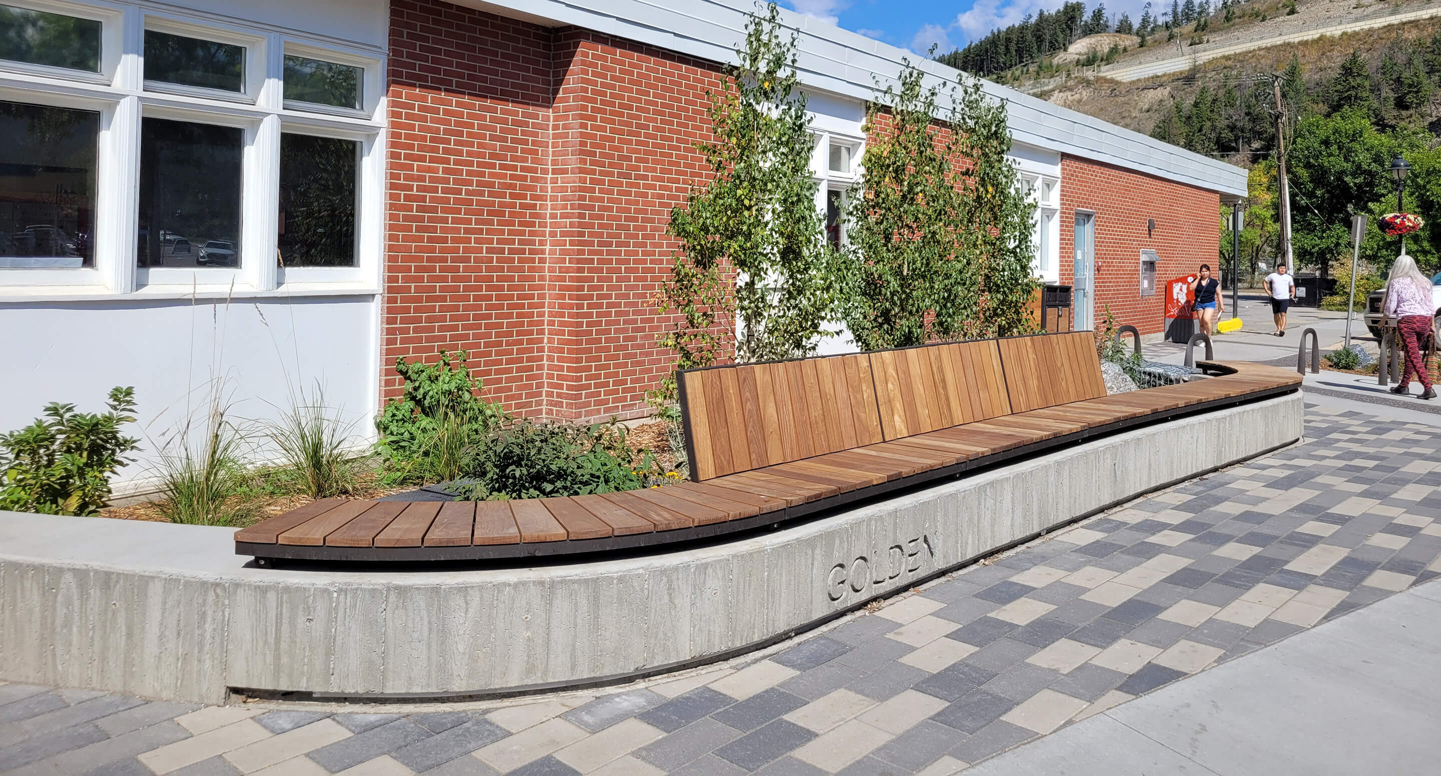 maglin-hero-img-ogden-wall-mount3 A long wooden Ogden bench with a backrest from the Ogden Collection is built into a curved concrete base in front of a red brick building with windows, small trees, and a patterned walkway.