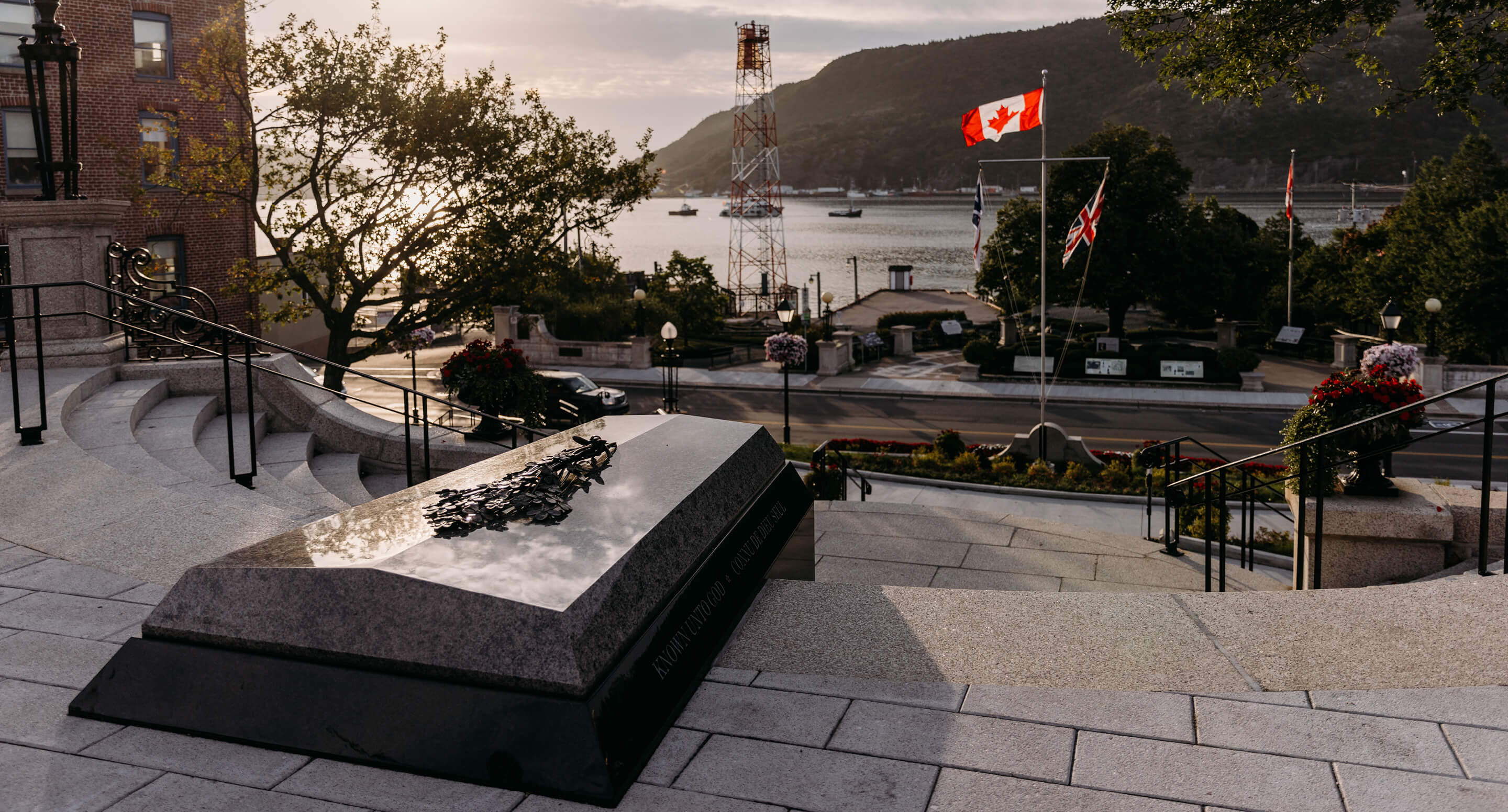 The Tomb of the Unknown Soldier, part of the National War Memorial in Newfoundland, stands on a set of steps overlooking a river, with Canadian and British flags and a sunset in the background.