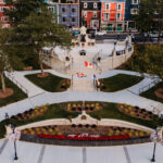 Aerial view of a landscaped public park with flowerbeds, paved walkways, flagpoles, statues—including the National War Memorial—trees, and colorful buildings in the background of Newfoundland.