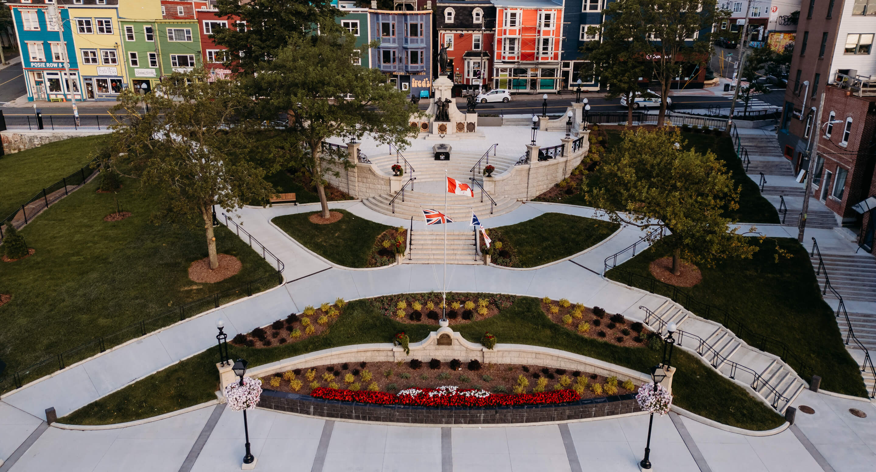 Aerial view of a landscaped public park with flowerbeds, paved walkways, flagpoles, statues—including the National War Memorial—trees, and colorful buildings in the background of Newfoundland.