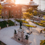A city square with the National War Memorial as its central monument, surrounded by trees, Newfoundland brick buildings, and a docked ship by the waterfront at sunset.