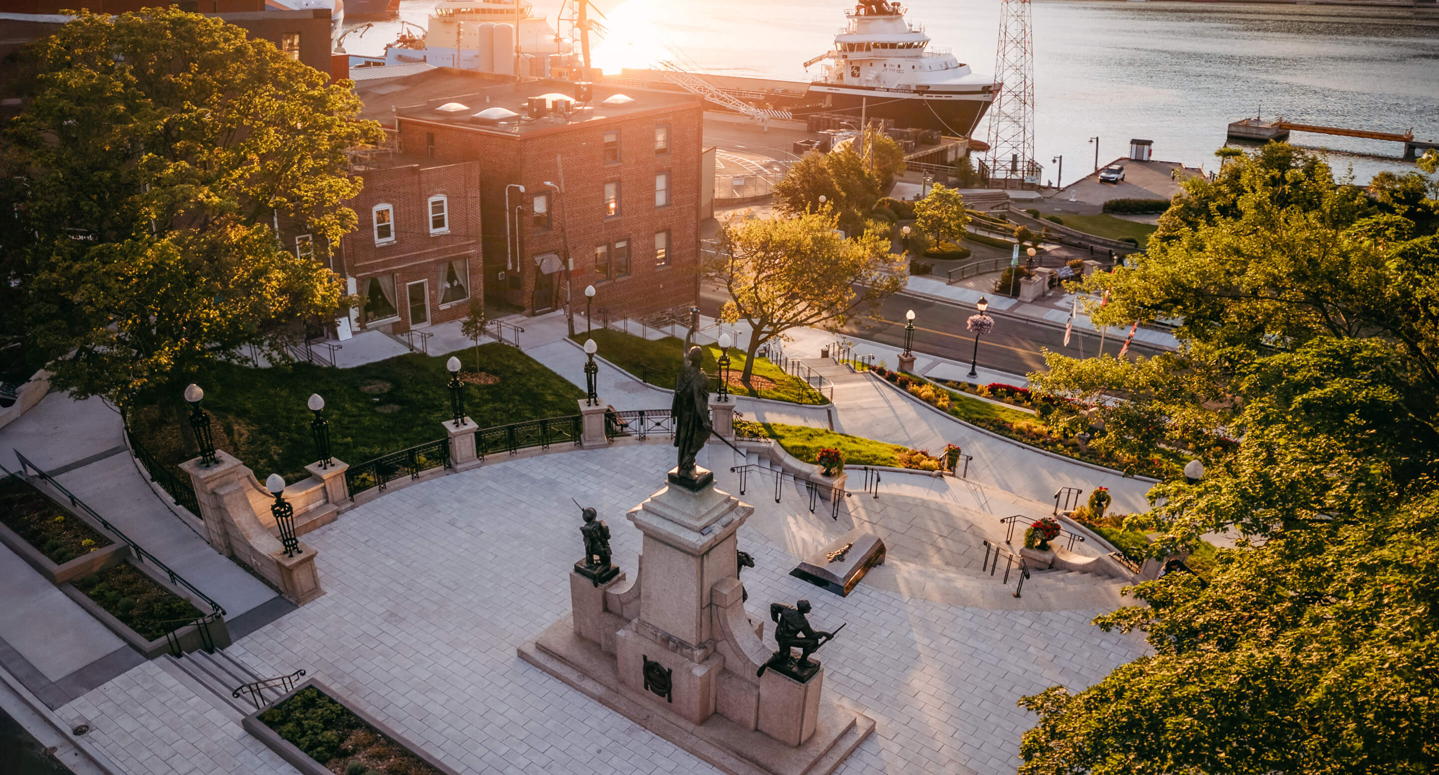 A city square with the National War Memorial as its central monument, surrounded by trees, Newfoundland brick buildings, and a docked ship by the waterfront at sunset.