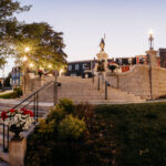Stone stairs lead up to the National War Memorial, surrounded by street lamps and flowers in a landscaped urban park at dusk.