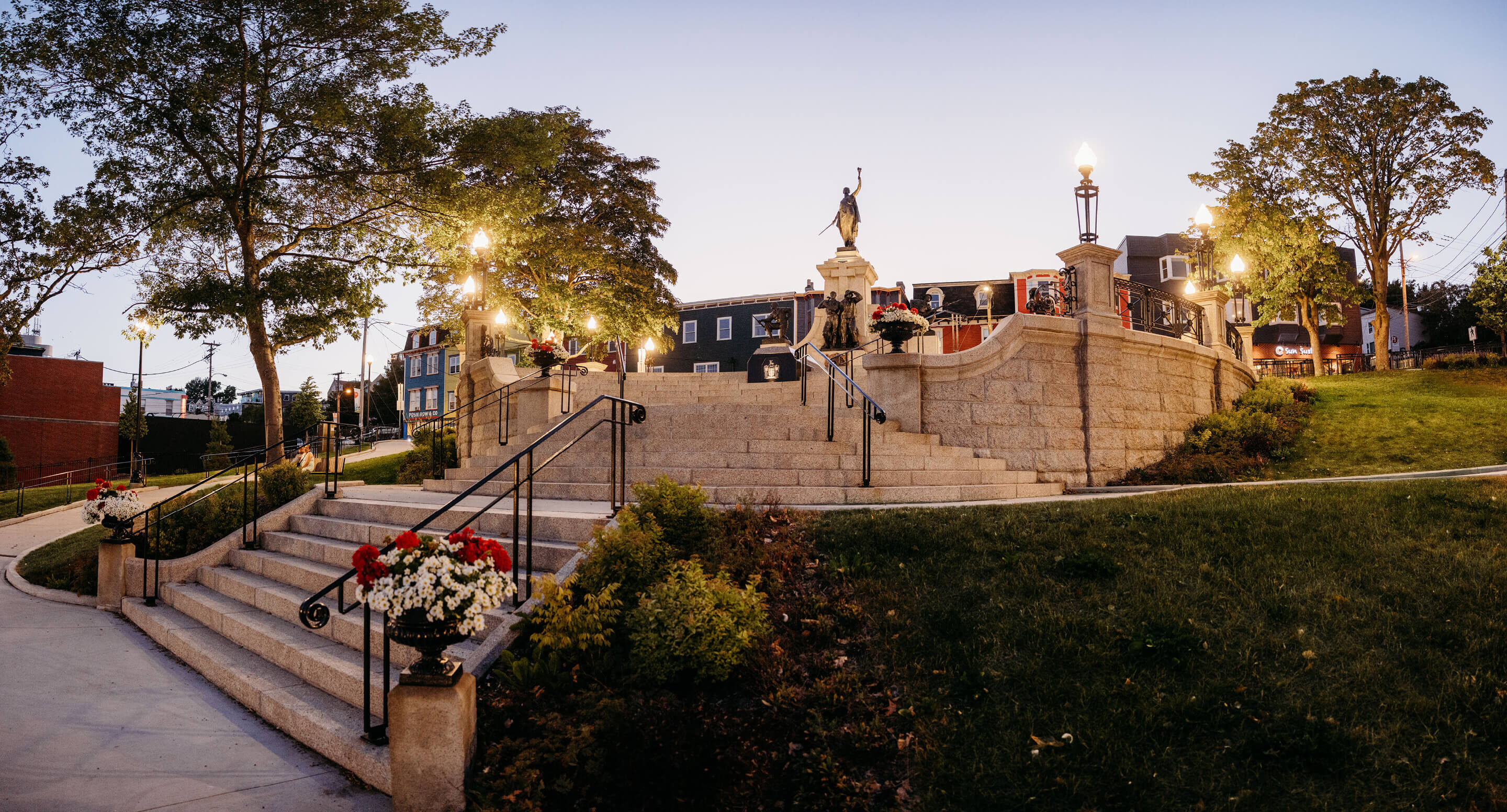 Stone stairs lead up to the National War Memorial, surrounded by street lamps and flowers in a landscaped urban park at dusk.