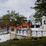 Stone monument with steps and railings in an urban park, surrounded by buildings, trees, streetlights, and flagpoles displaying Canadian and British flags—this is the Newfoundland National War Memorial.