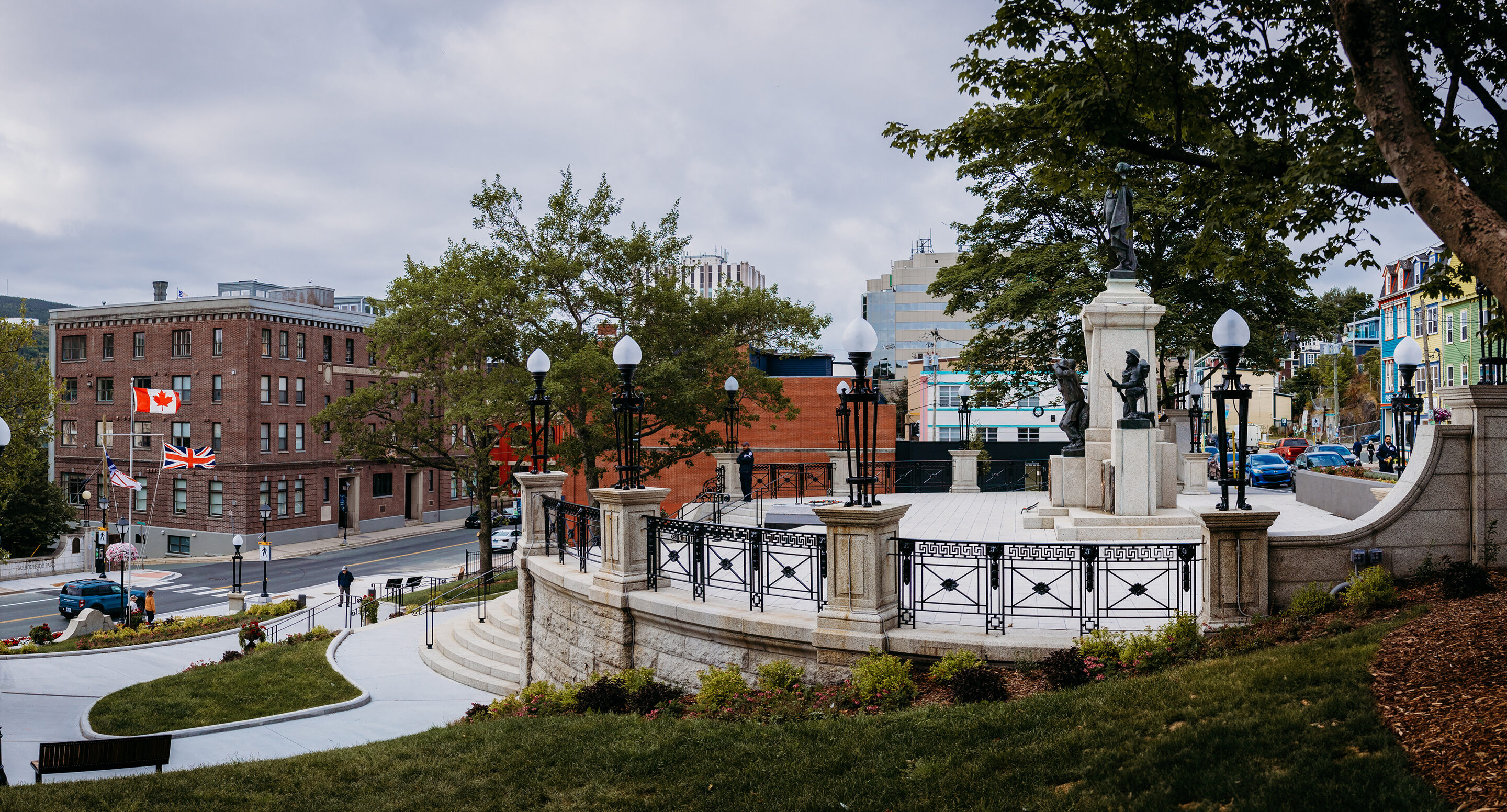 Stone monument with steps and railings in an urban park, surrounded by buildings, trees, streetlights, and flagpoles displaying Canadian and British flags—this is the Newfoundland National War Memorial.