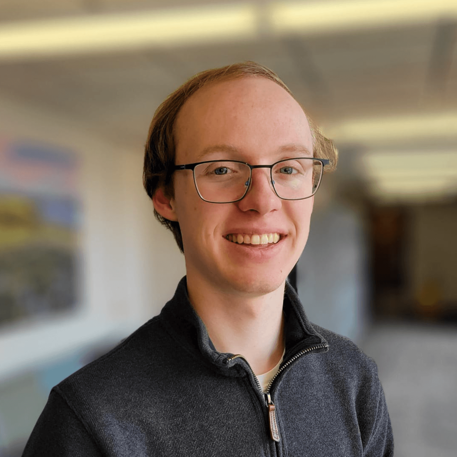 A young person with glasses and short brown hair smiles at the camera, wearing a dark zip-up sweater in an indoor setting with blurred background.