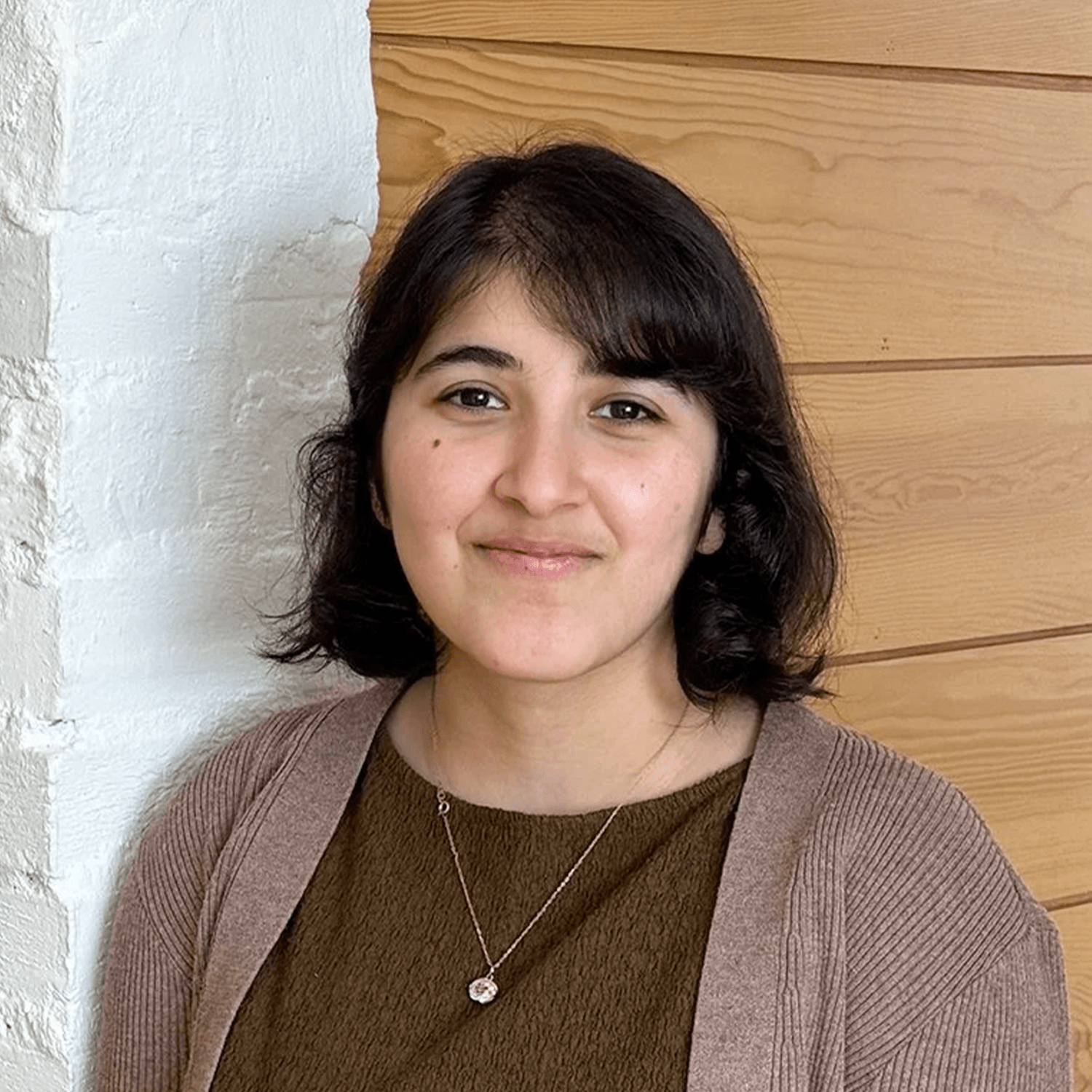 A person with short dark hair wearing a brown top, a light brown cardigan, and a necklace stands indoors in front of a white brick wall and wooden paneling.