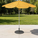 A large, yellow, flower-shaped ROMA umbrella stands on a paved square in a green park with trees in the background.