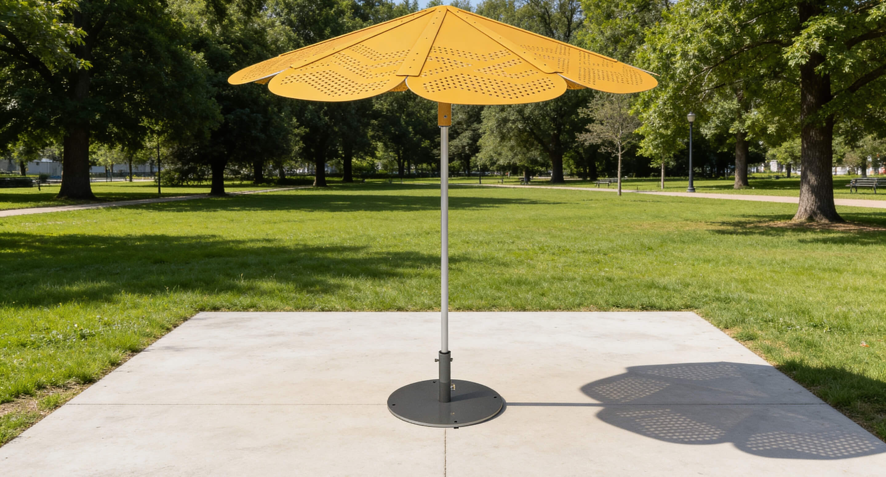 A large, yellow, flower-shaped ROMA umbrella stands on a paved square in a green park with trees in the background.