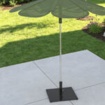 Reseda green ROMA sun shade and base with a leaf-like design stands on a concrete patio in a backyard with grass, flower beds, and a house wall.