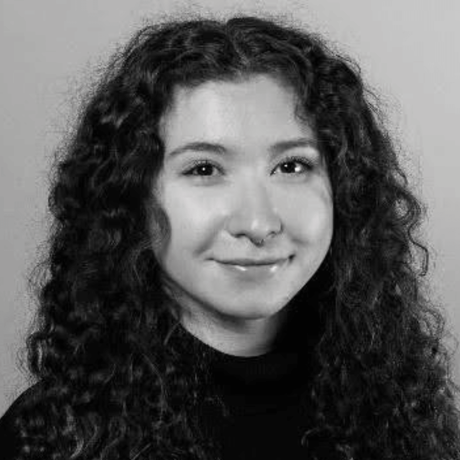 A young woman with long curly hair wearing a dark top, smiling slightly, in a black and white portrait against a plain background.