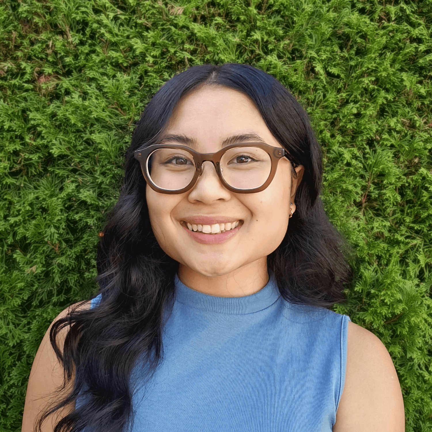 A woman with long dark hair and glasses smiles at the camera, standing in front of a green leafy background and wearing a sleeveless blue top.