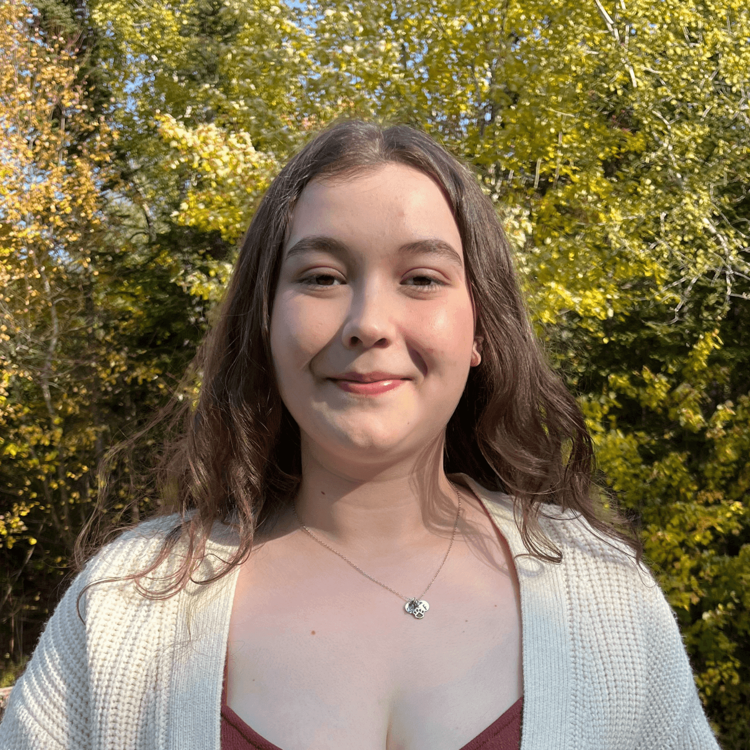A person with long brown hair stands outdoors, wearing a cream sweater and a necklace, with trees and sunlight in the background.