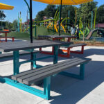 Picnic tables with attached benches and yellow umbrellas on a concrete area in Shoreview Commons, with a colorful playground and people enjoying the day in the background in Shoreview, Minnesota.