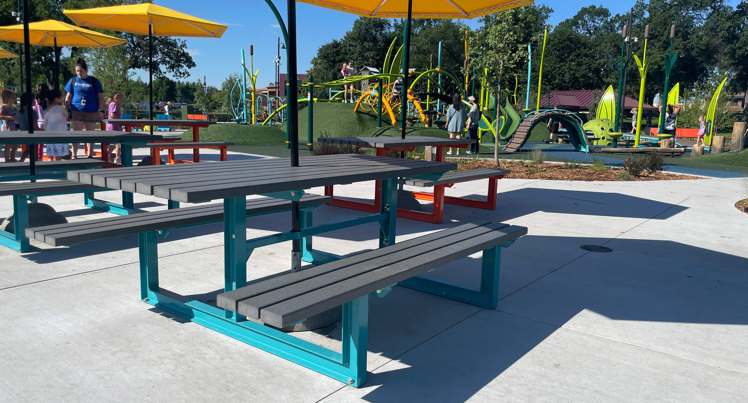 Picnic tables with attached benches and yellow umbrellas on a concrete area in Shoreview Commons, with a colorful playground and people enjoying the day in the background in Shoreview, Minnesota.