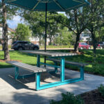 A turquoise picnic table with attached benches and a large umbrella sits on a concrete patio at Shoreview Commons in Minnesota, surrounded by grass, trees, and parked cars.
