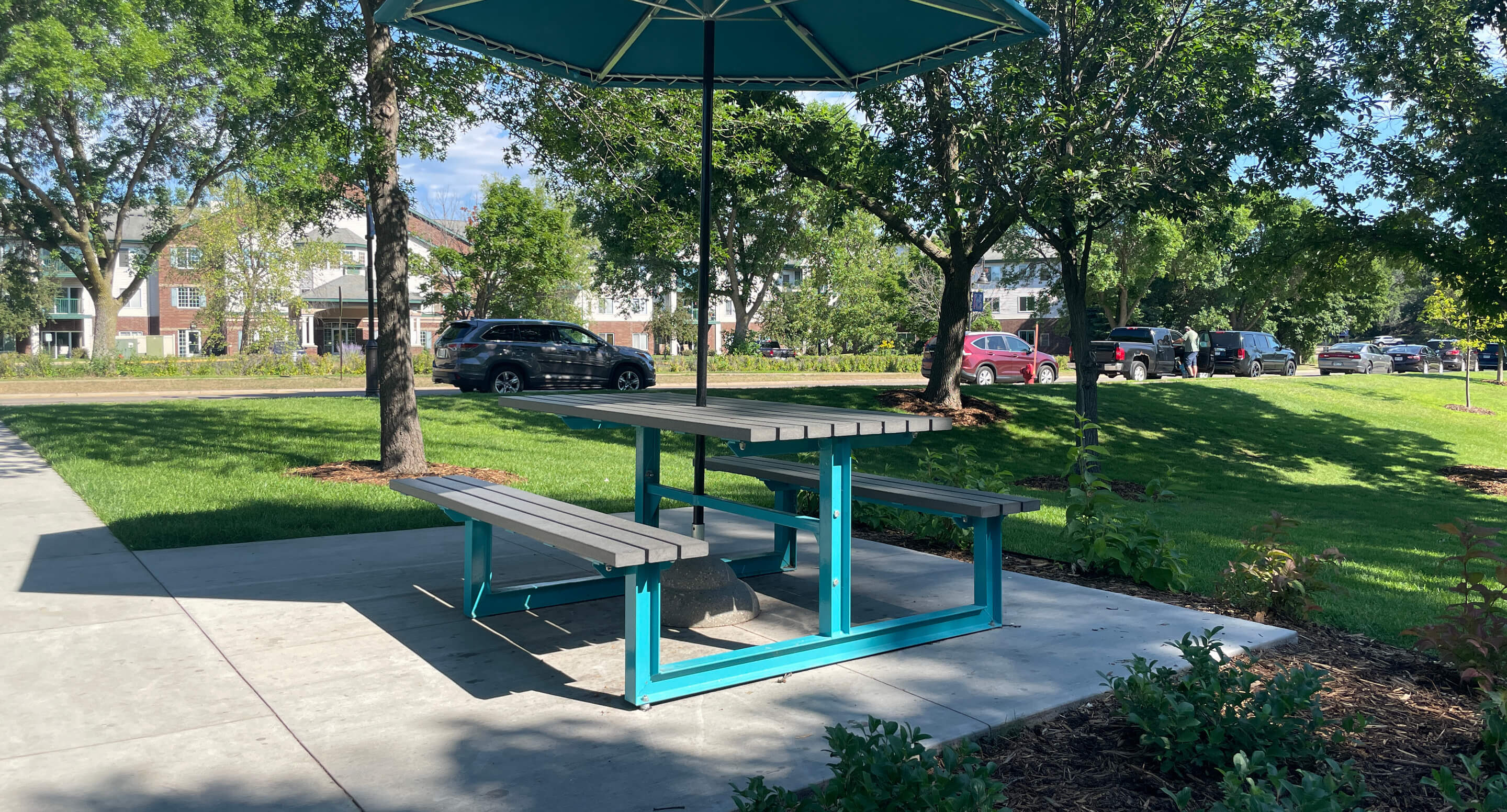 A turquoise picnic table with attached benches and a large umbrella sits on a concrete patio at Shoreview Commons in Minnesota, surrounded by grass, trees, and parked cars.