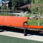 A long orange bench with metal armrests is situated on a path in Shoreview Commons, a landscaped park area in Shoreview, Minnesota, with trees, plants, and playground equipment in the background.