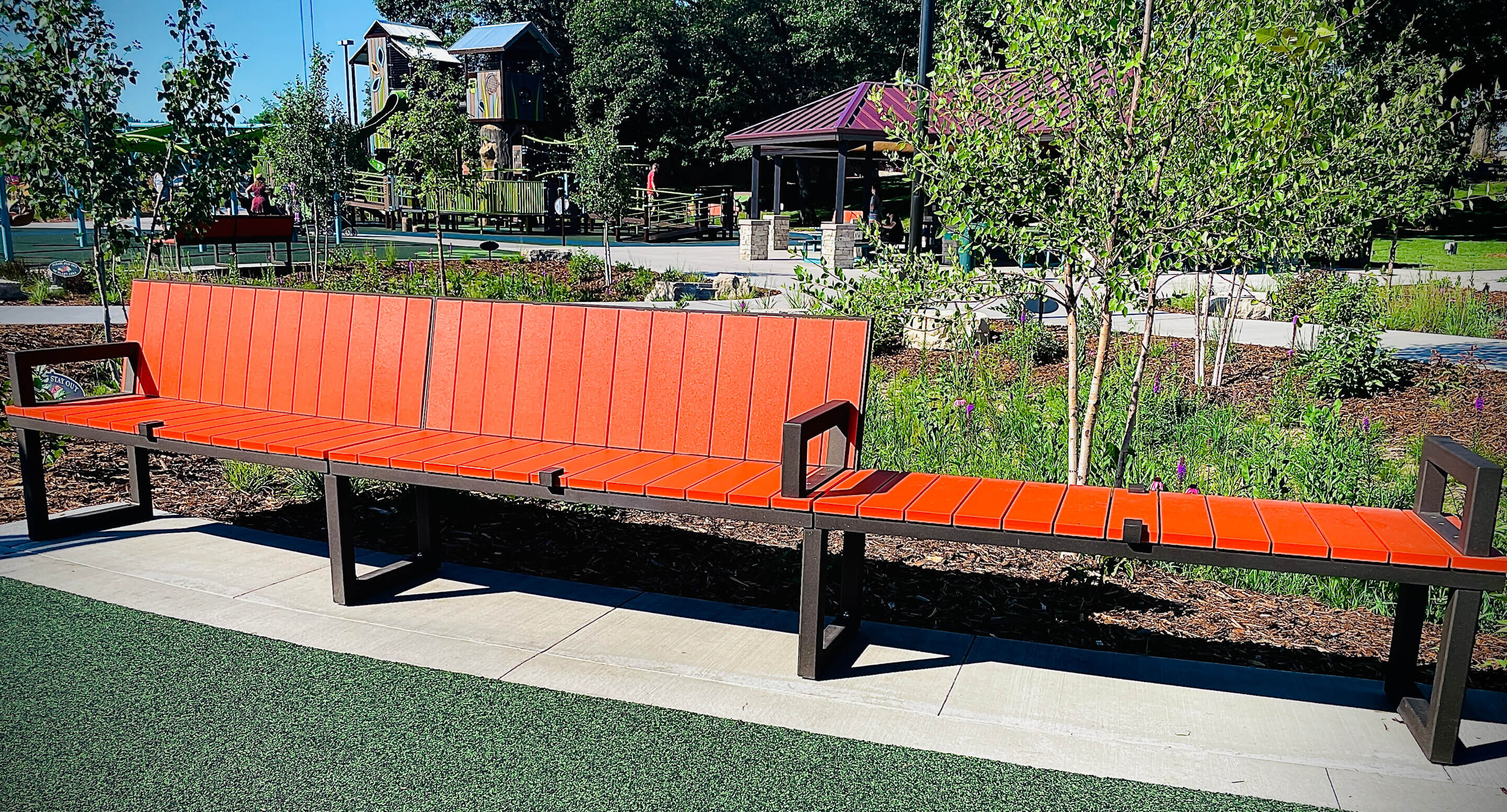 A long orange bench with metal armrests is situated on a path in Shoreview Commons, a landscaped park area in Shoreview, Minnesota, with trees, plants, and playground equipment in the background.
