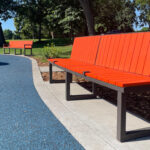 Two orange Ogden benches with bronze metal frames are placed along a curved path in Shoreview Commons, a shaded park area in Shoreview, Minnesota, with trees and blue rubberized ground.