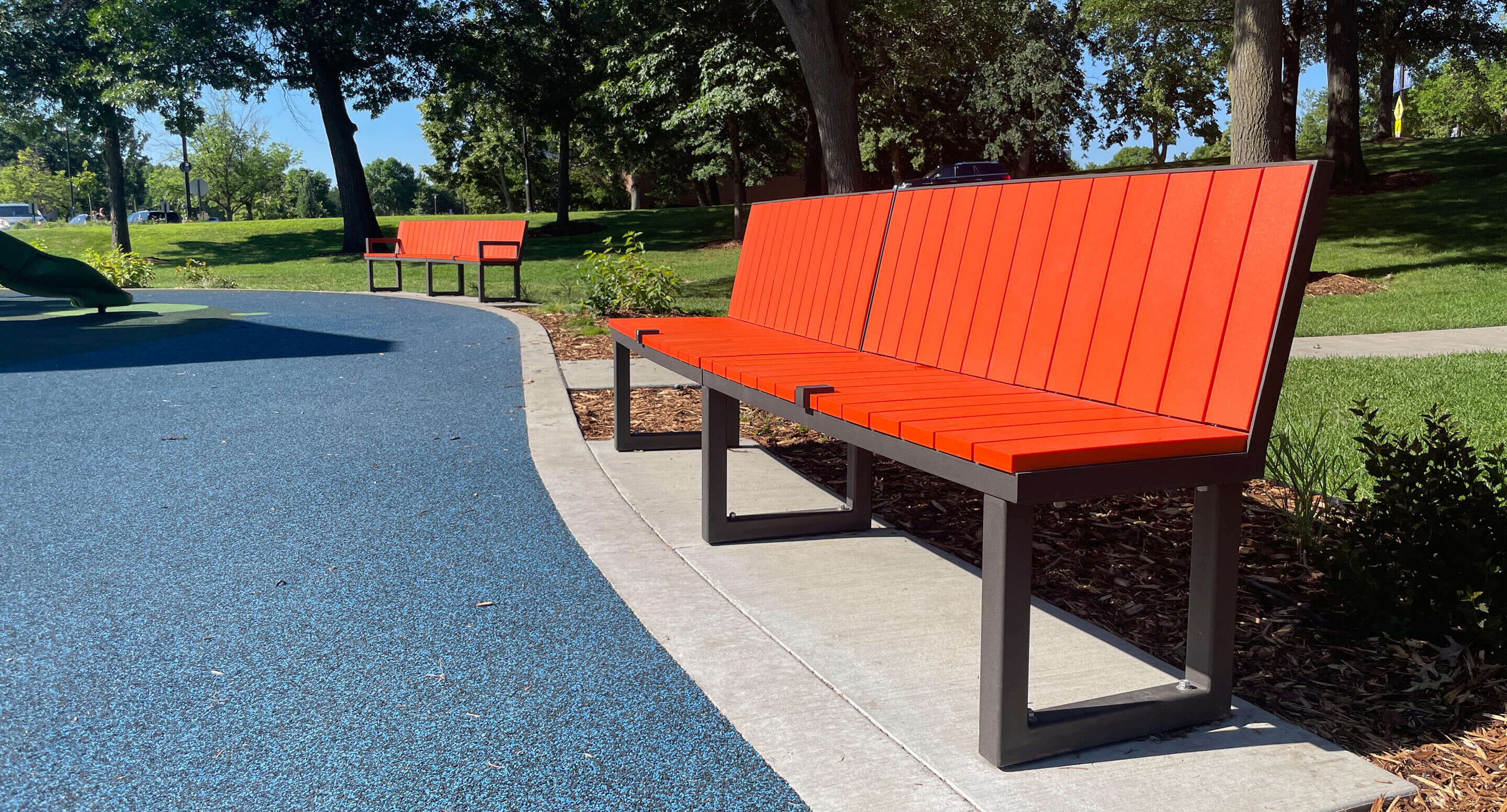 Two orange Ogden benches with bronze metal frames are placed along a curved path in Shoreview Commons, a shaded park area in Shoreview, Minnesota, with trees and blue rubberized ground.