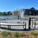 Circular Ogden benches surround a paved area in front of a pond with a central fountain at Shoreview Commons, beautifully landscaped with plants and a building in the background in Shoreview, Minnesota.