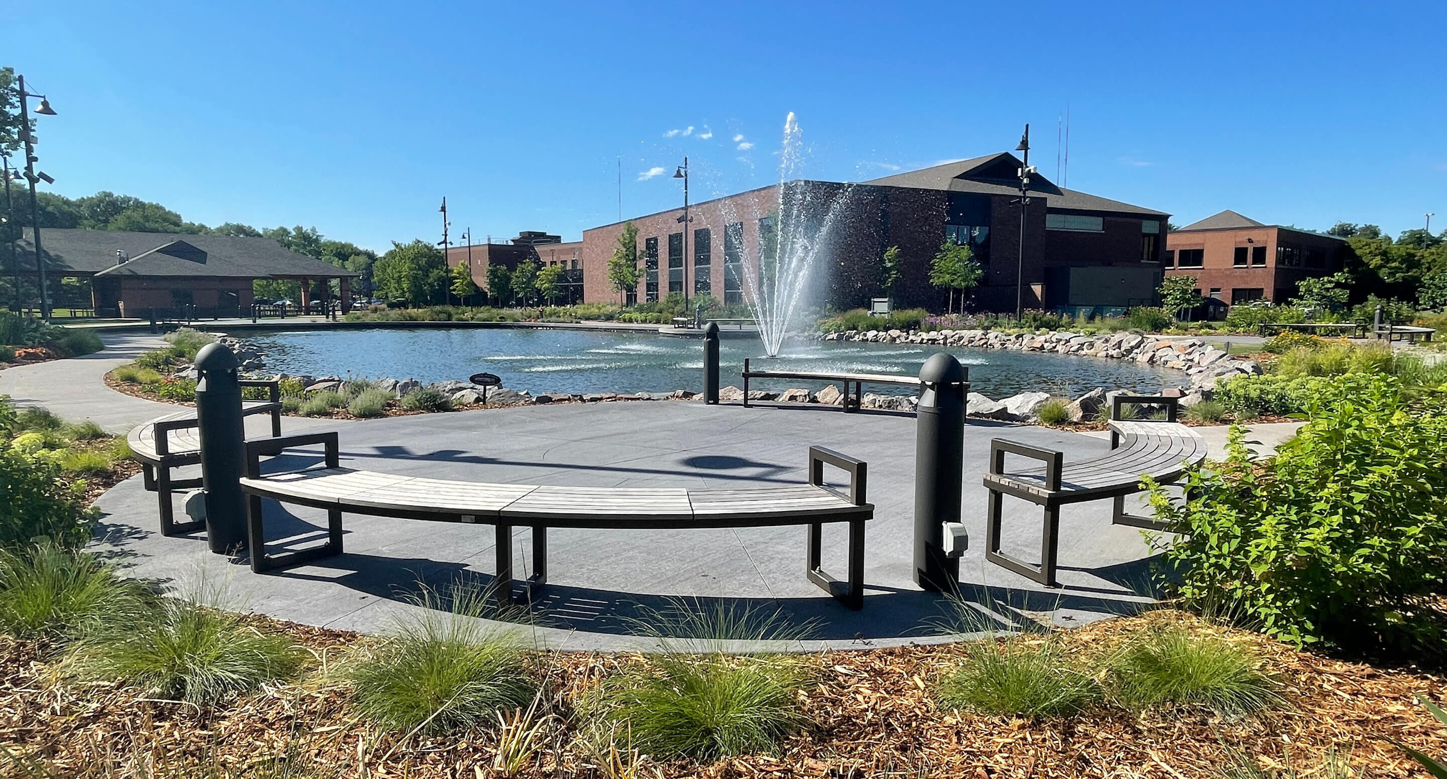 Circular Ogden benches surround a paved area in front of a pond with a central fountain at Shoreview Commons, beautifully landscaped with plants and a building in the background in Shoreview, Minnesota.