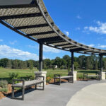 Curved metal pergola with patterned roof, Ogden benches, and stone pillars along a walkway in Shoreview Commons park, Shoreview, Minnesota, surrounded by grass, trees, and blue sky.