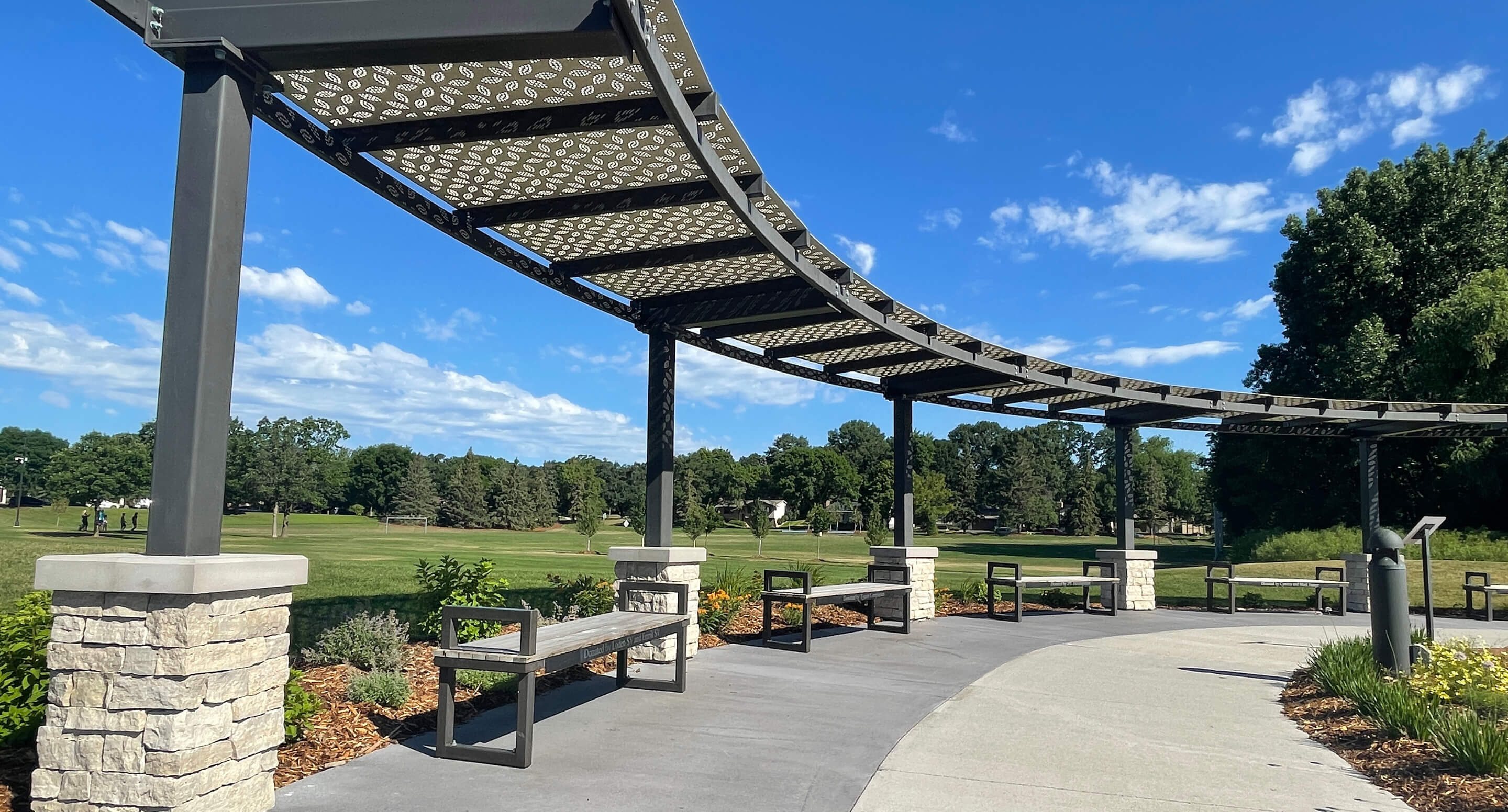 Curved metal pergola with patterned roof, Ogden benches, and stone pillars along a walkway in Shoreview Commons park, Shoreview, Minnesota, surrounded by grass, trees, and blue sky.