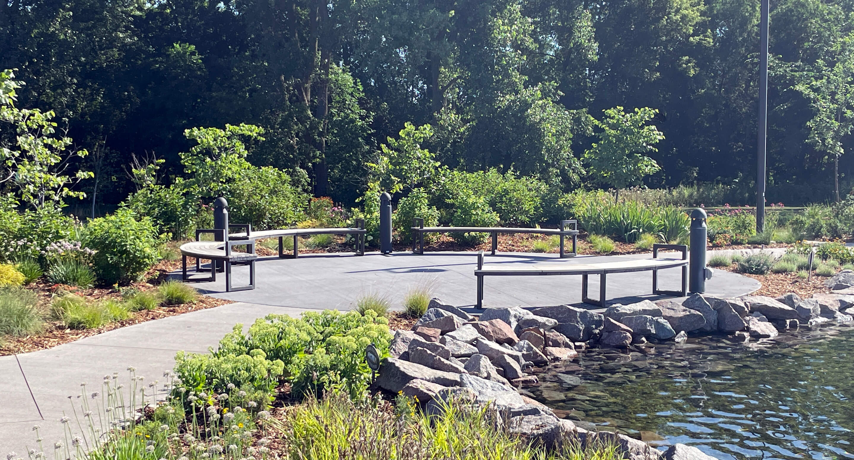 Curved Ogden Benches are situated on a paved area by a pond, surrounded by greenery and trees in the landscaped Shoreview Commons park in Shoreview, Minnesota.
