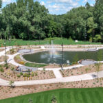 Shoreview Commons in Minnesota features a landscaped park with a pond and central fountain, walking paths, benches, young trees, and surrounding greenery under a partly cloudy sky.