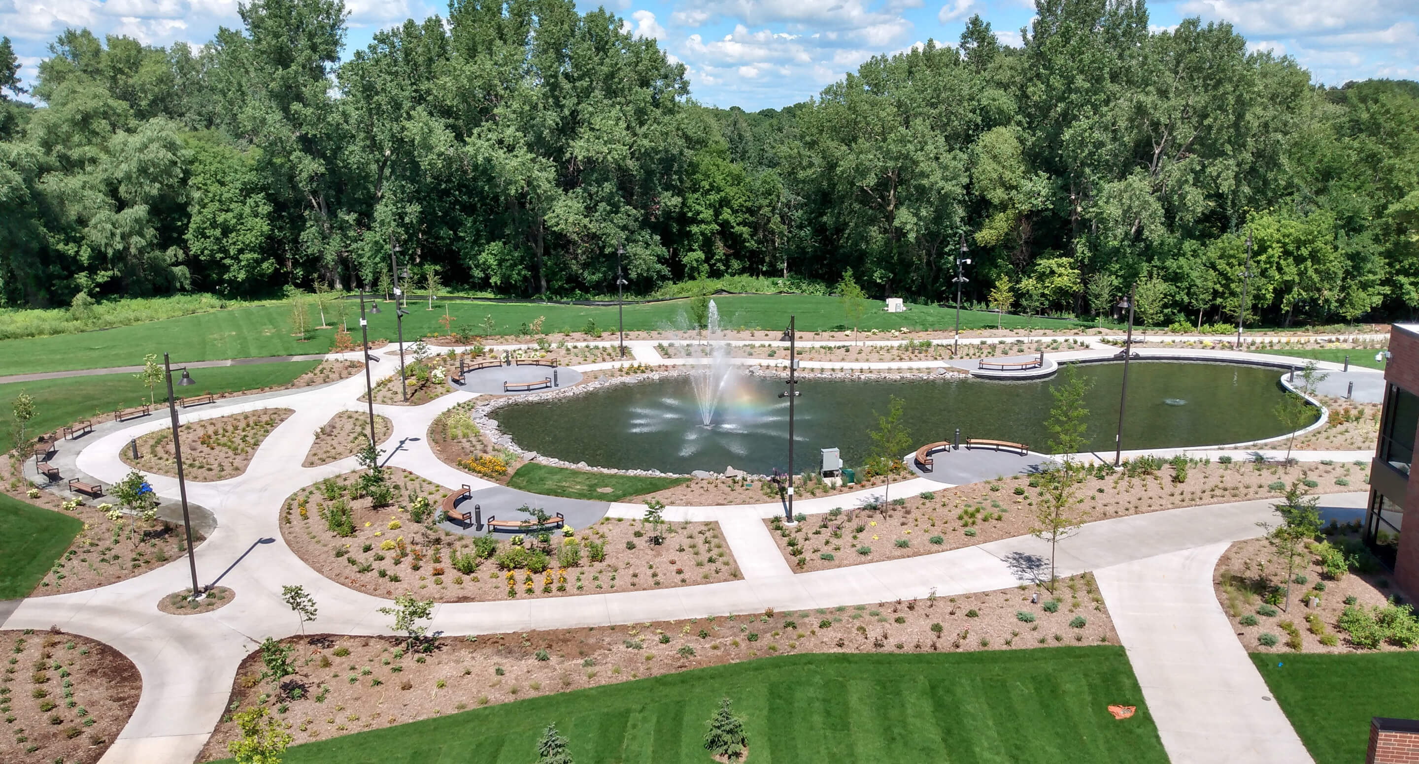 Shoreview Commons in Minnesota features a landscaped park with a pond and central fountain, walking paths, benches, young trees, and surrounding greenery under a partly cloudy sky.