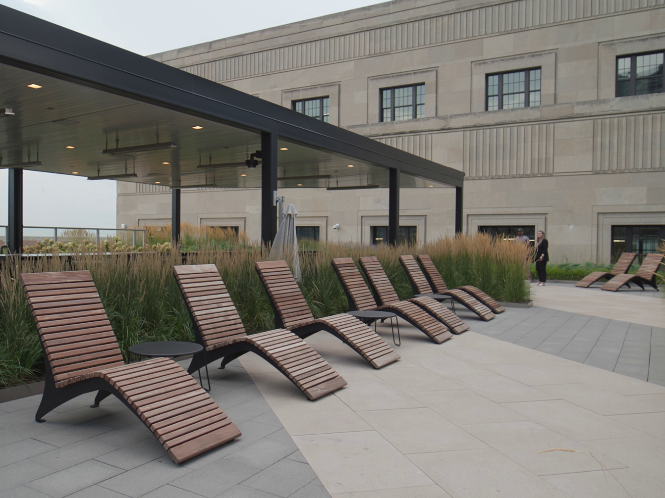 A row of wooden lounge chairs is arranged on a rooftop patio next to tall grasses, with a modern pergola and a large stone building in the background.