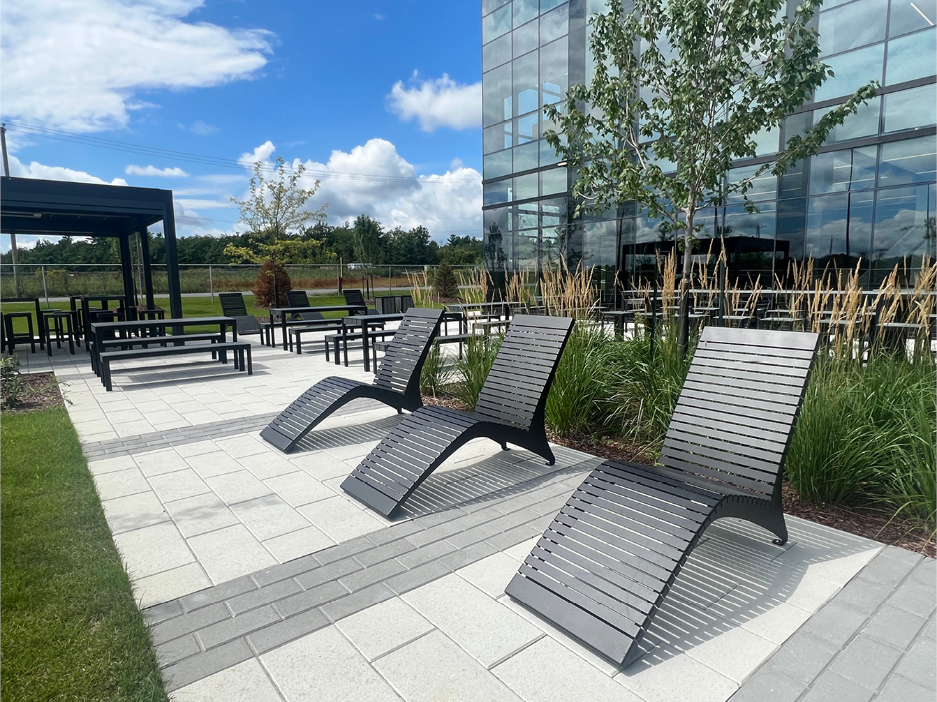Outdoor patio with black lounge chairs, tables, and benches on a paved surface beside a modern glass building, surrounded by grass, plants, and trees under a partly cloudy sky.