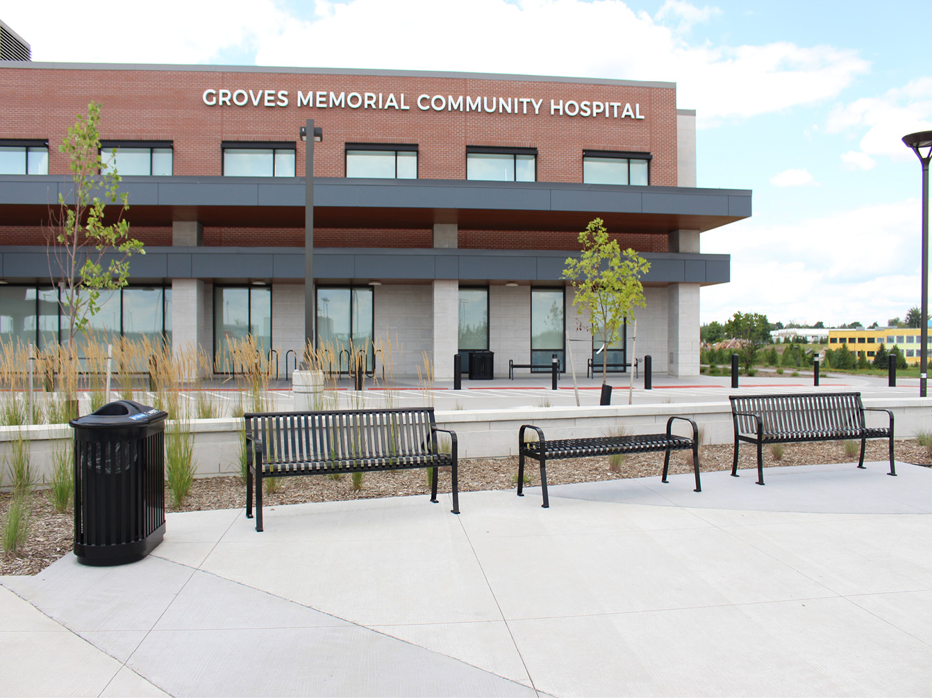 Exterior view of Groves Memorial Community Hospital with outdoor areas featuring metal benches, small trees, and a trash bin in the foreground.