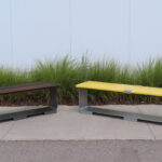 Two modern metal benches, one brown and one yellow, are placed on a sidewalk in front of tall green grass and a white wall—perfect seating for outdoor areas.