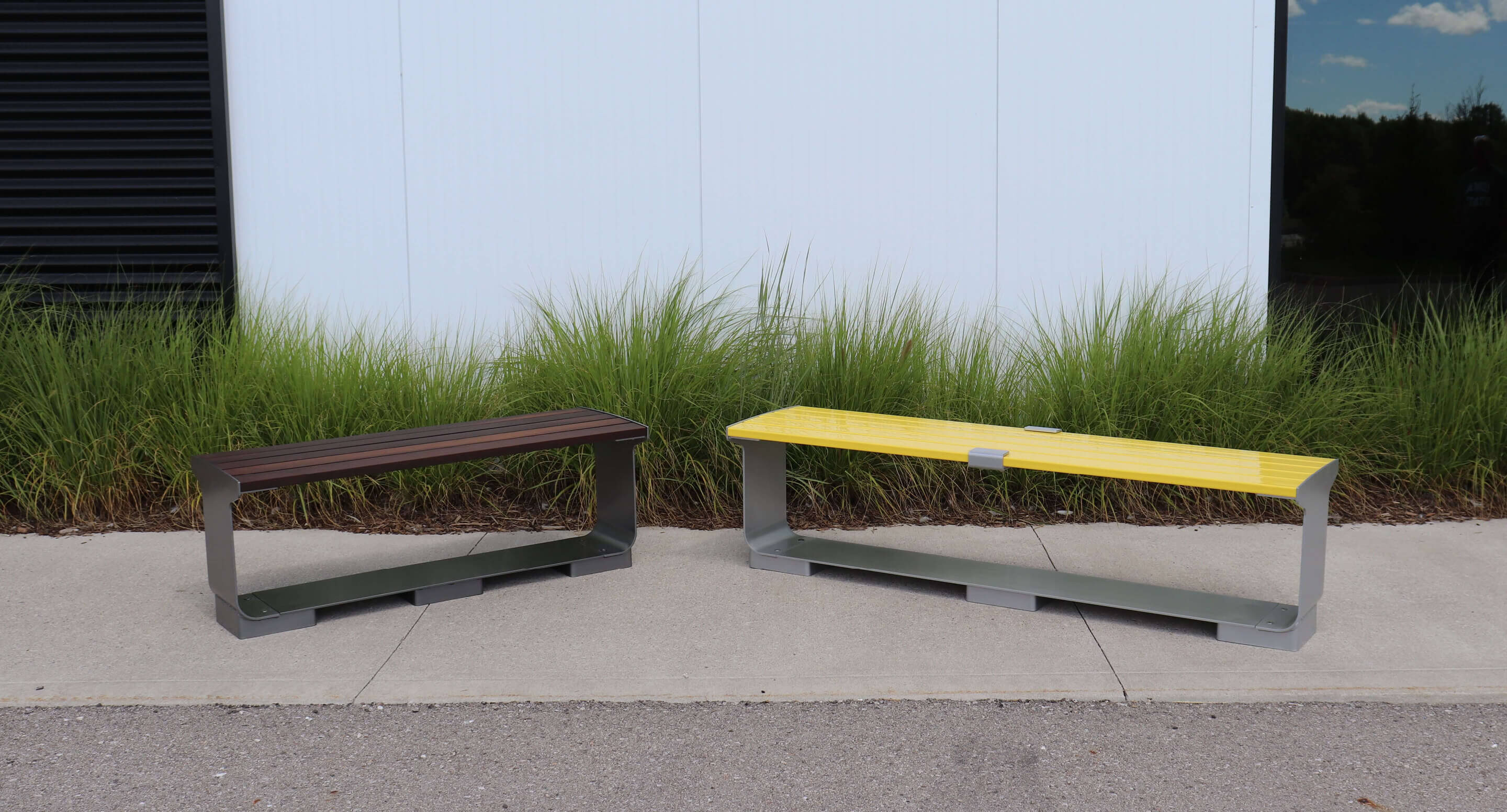 Two modern metal benches, one brown and one yellow, are placed on a sidewalk in front of tall green grass and a white wall—perfect seating for outdoor areas.
