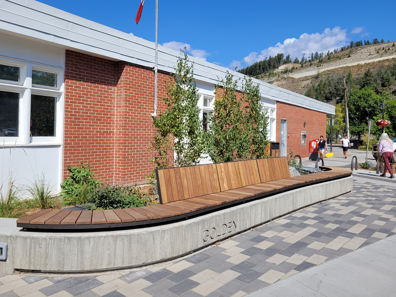 A modern wooden bench labeled "GOLDEN" sits outside a red-brick building with large windows, enhancing outdoor areas where people stroll nearby under a blue sky.