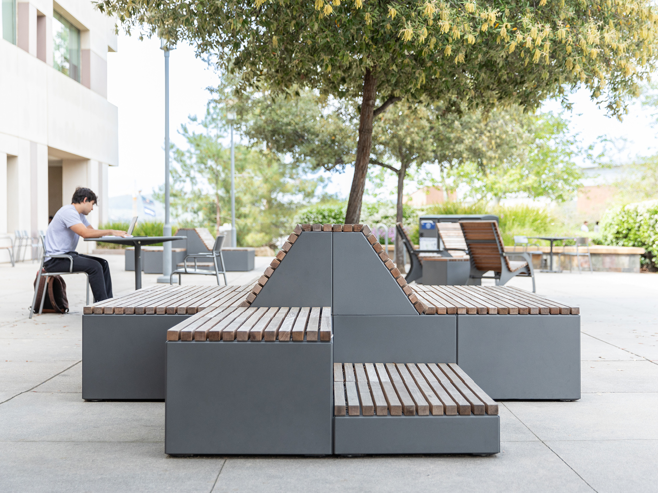 Modern outdoor seating - Maglin's Pixel Collection - with wooden slats and steel base, shaded by a tree; a person sits at a table nearby with a bag on the ground.