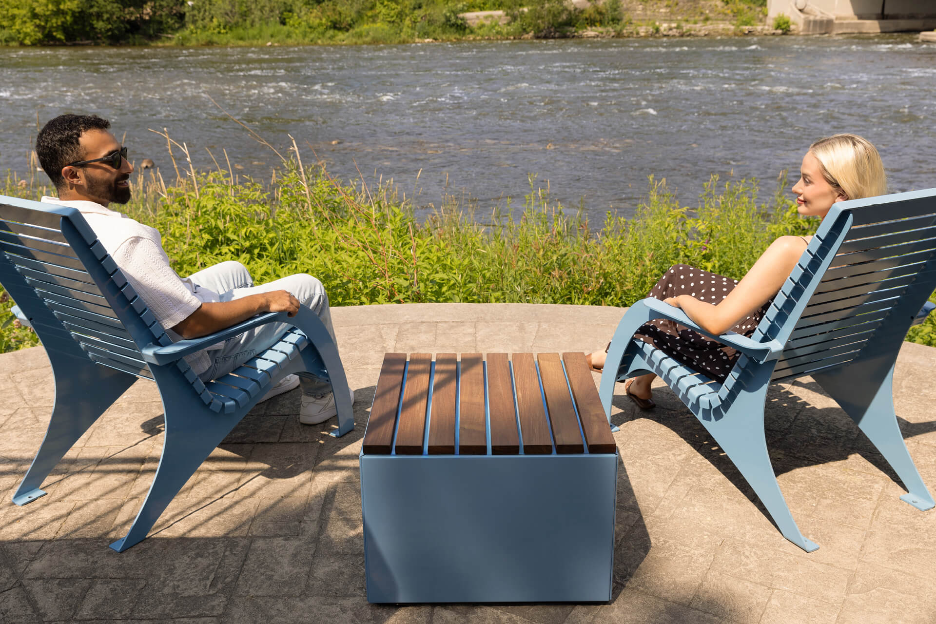 Two people sit in blue Maglin site furniture 720 chairs facing a river, separated by a small Pixel table, on a stone patio with green plants in the background.