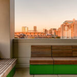 Outdoor seating area with wooden Pixel Collection benches and green bases, overlooking a city skyline with tall buildings during sunset.