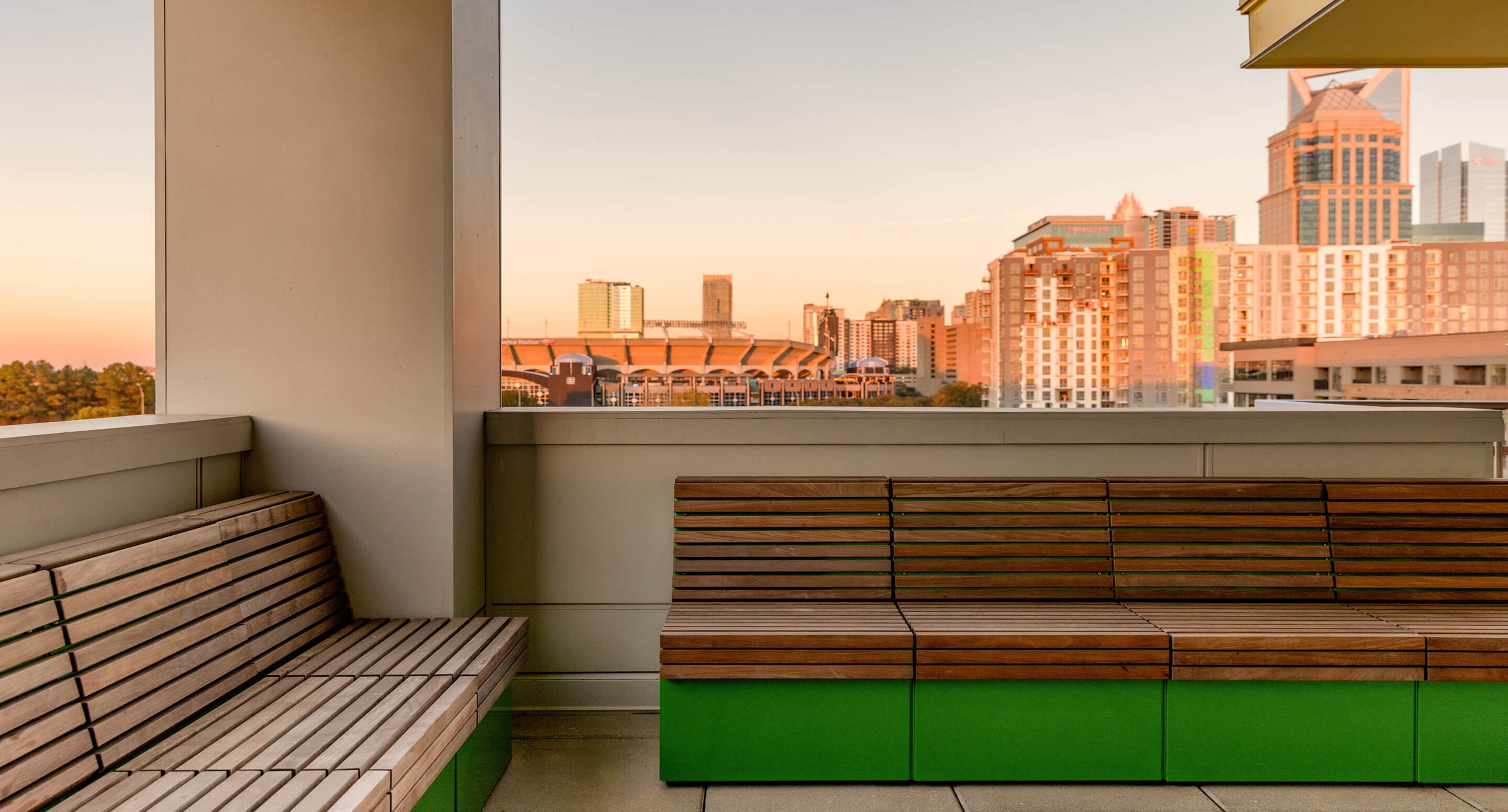 Outdoor seating area with wooden Pixel Collection benches and green bases, overlooking a city skyline with tall buildings during sunset.