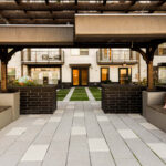 Modern apartment courtyard at Link Apartments in Charlotte NC, featuring wooden benches, pillows, a paved walkway, an inviting pergola overhead, and a view of balconies and doors lining Mint Street in the background.