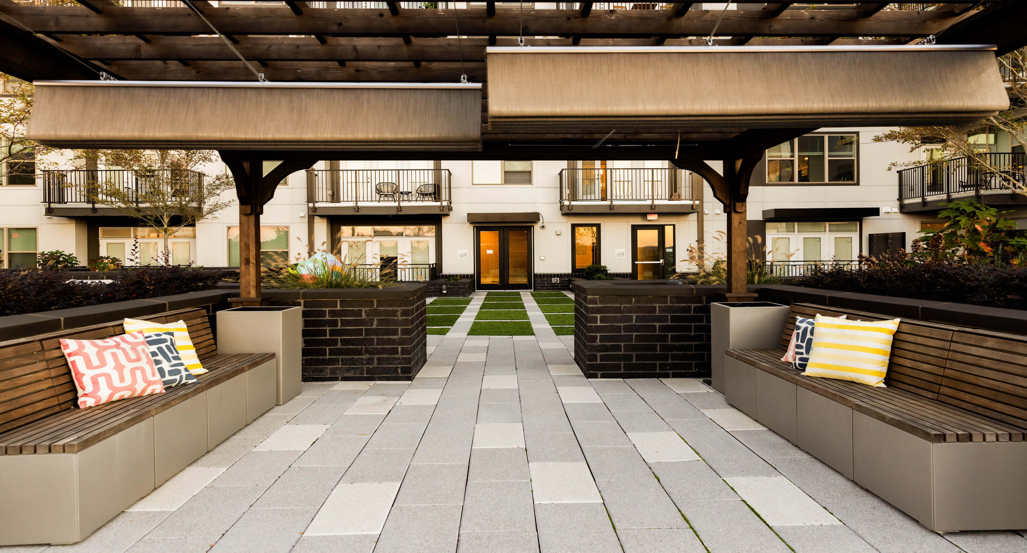 Modern apartment courtyard at Link Apartments in Charlotte NC, featuring wooden benches, pillows, a paved walkway, an inviting pergola overhead, and a view of balconies and doors lining Mint Street in the background.